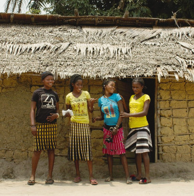 A photo of four women standing in front of a cob/mud house built with a palm leaf roof