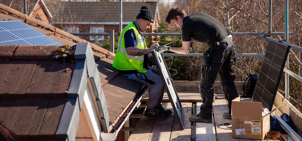 Two men fitting solar panels to the roof of a building
