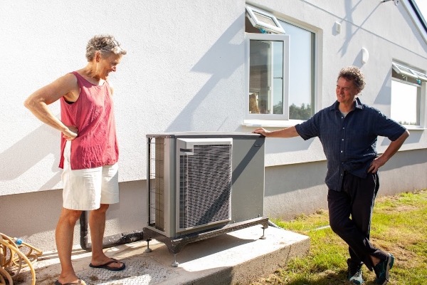 A man and a woman standing in front of a heat pump unit next to their home