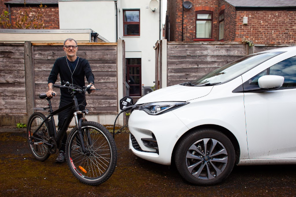 A photo of a man with an electric bicycle next to an electric vehicle in front of a terraced house with external wall insualtion.