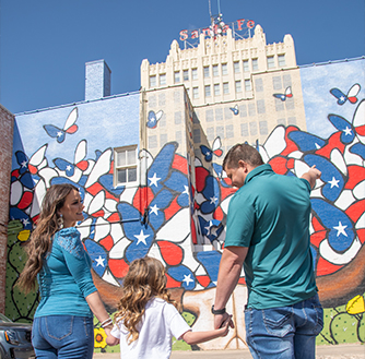 A family with a child admire a red, white, and blue mural painted on a building wall.